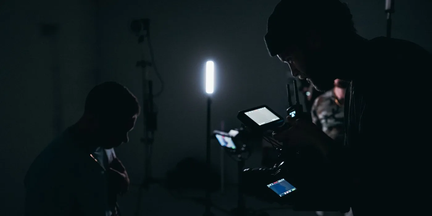 A camera operator with a rigged cinema camera and an external monitor films a seated subject in a darkened studio lit by a single tube light.